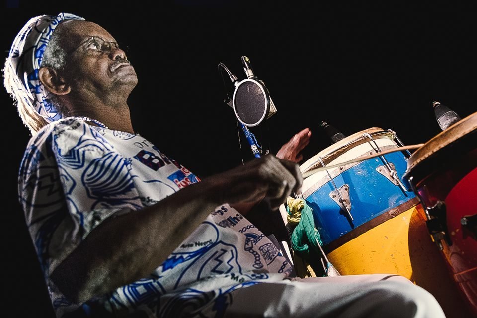 Foto colorida do mestre Môa do Katendê. Ele está de roupa branca com detalhes em azul e tocando instrumentos de percussão - Metrópoles