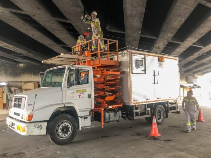 foto colorida, operários da Prefeitura trabalham na manutenção de viaduto. Eles estao num caminhão na parte de baixo do viaduto