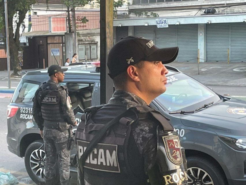 Dois policiais militares fardados e com colete à prova de balas; estão de pé, perto de viatura, em rua do Guarujá, foto colorida
