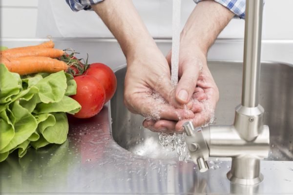 Foto colorida de pessoa branca lavando as mãos em uma pia. Ao lado, há cenouras, tomates e verduras - Metrópoles