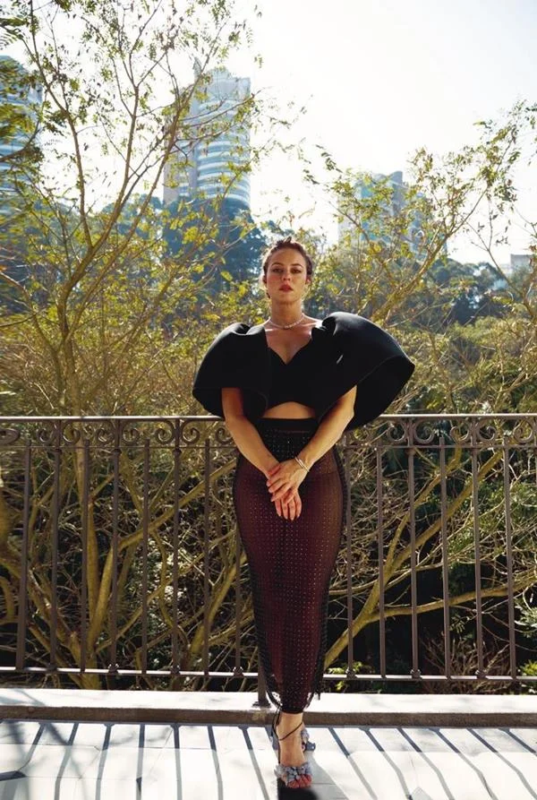 Mulher posando para foto em frente a uma sacada. Ela est&aacute; com os bra&ccedil;os cruzados e olhando para a c&acirc;mera - Metr&oacute;poles