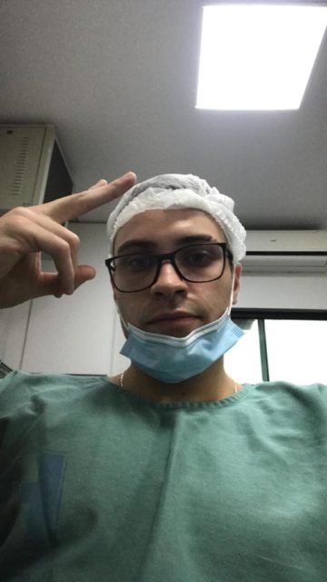 imagem colorida mostra homem loiro de olhos verdes, pele branca, óculos de grau com armação preta usando camiseta verde e touca cirurgica branca - Metrópoles