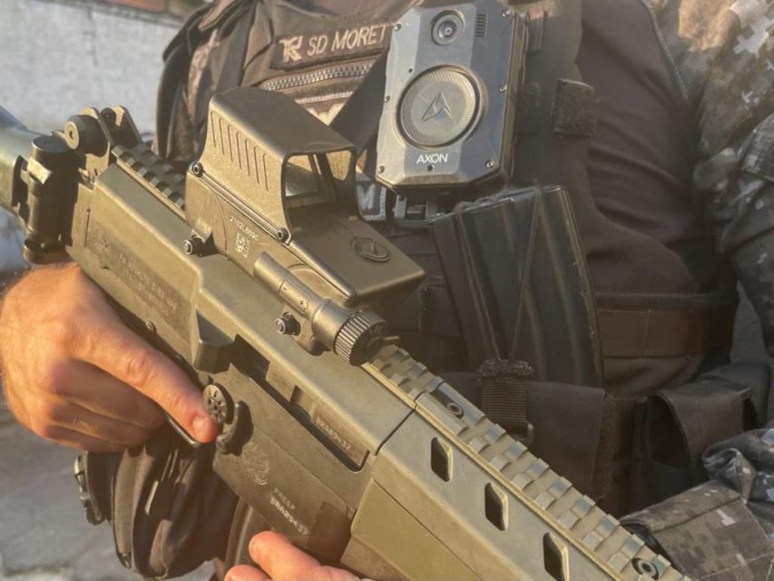 Foto colorida mostra policial militar segurando arma de fogo