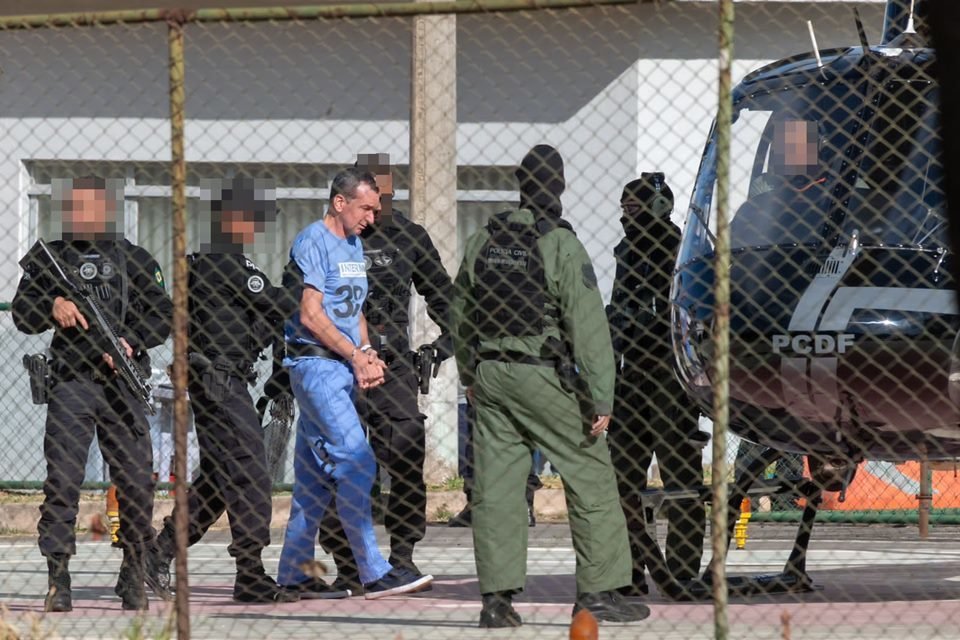 Marcola, homem branco, magro de cabelos raspados, usa uniforme azul e é escoltado por cinco militares, de uniformes nas cores preta e verde; ele é encaminhado para um helicóptero preto; os rostos dos policiais aparecem borrados