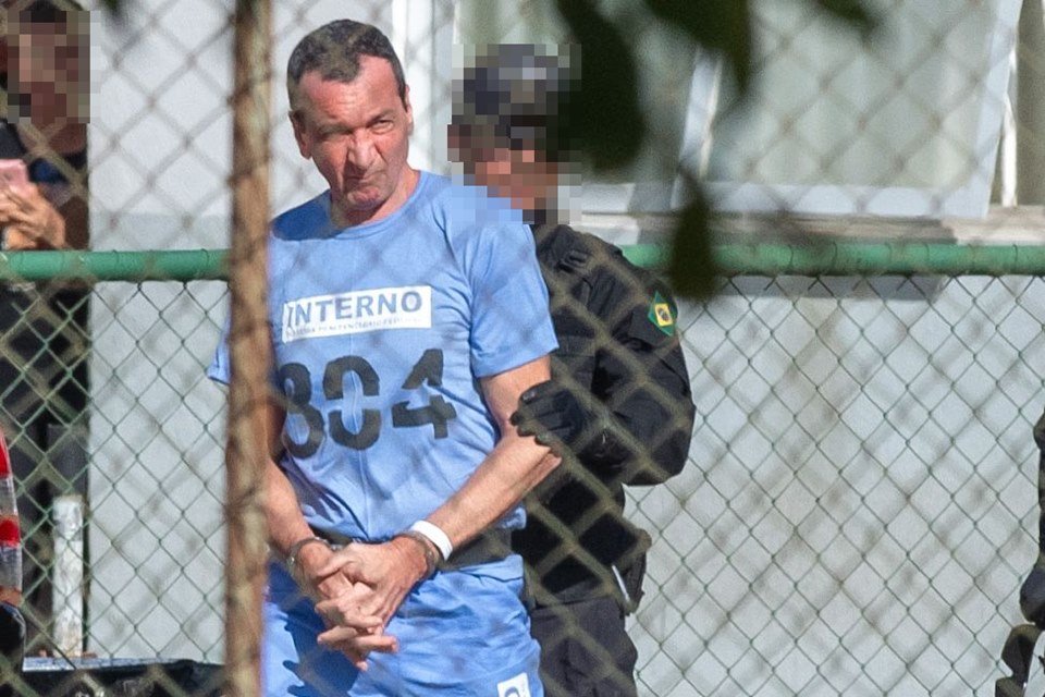 Marcola, homem branco, magro, de cabelos curtos e escuros, veste uniforme azul, usado na prisão; ele é escoltado por um policial de uniforme preto e com rosto borrado