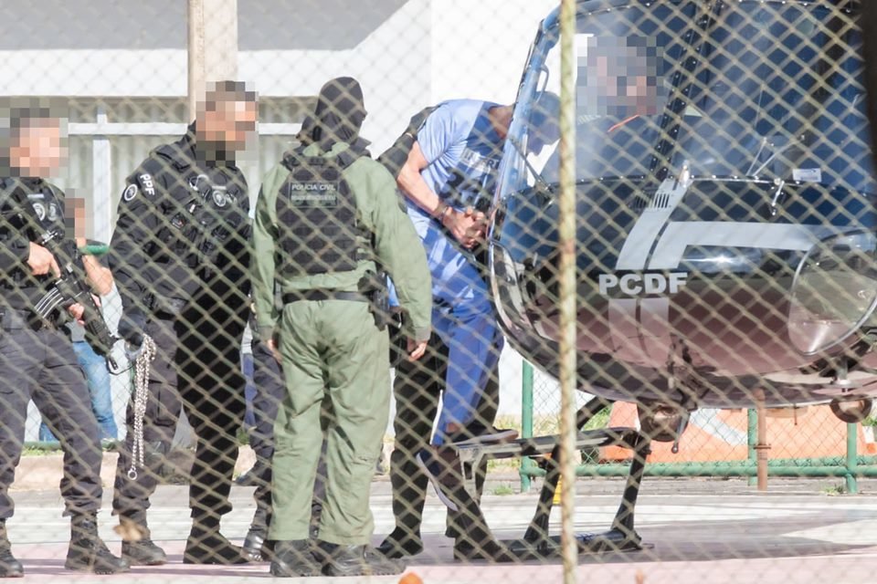 Marcola, homem branco, magro, de cabelos curtos e escuros, veste uniforme azul, usado na prisão; ele é escoltado por policiais de uniformes pretos e verdes e com rostos borrados; os policiais colocam Marcola em um helicóptero