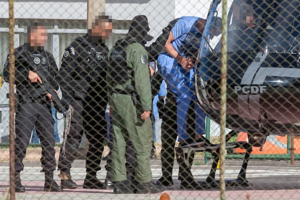Marcola, homem branco, magro, de cabelos curtos e escuros, veste uniforme azul, usado na prisão; ele é escoltado por policiais de uniformes pretos e verdes e com rostos borrados; os policiais conduzem Marcola para um helicóptero