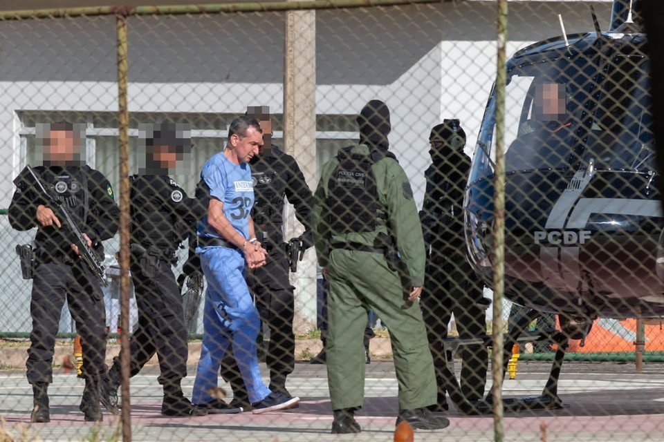 Marcola, homem branco, magro, de cabelos curtos e escuros, veste uniforme azul, usado na prisão; ele é escoltado por policiais de uniformes pretos e verdes e com rostos borrados; os policiais conduzem Marcola para um helicóptero