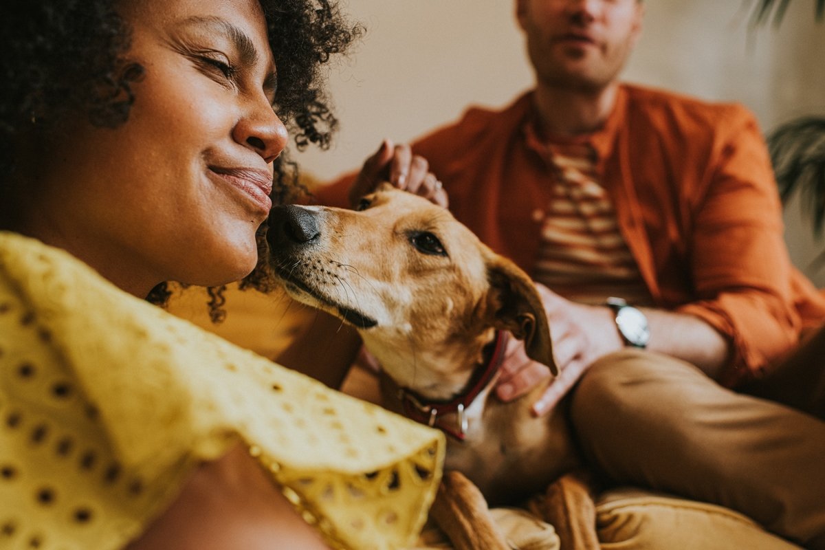 Foto colorida. Imagem mostra mulher negra sentada do lado esquerdo recebendo carinho do cachorrinho. No fundo, tem um homem sentado - Metr&oacute;poles