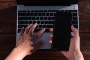 Fotografia colorida mostrando uma mão segurando celular e outra mão digitando em computador-Metrópoles