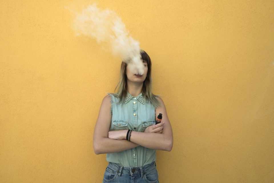 Jovem mulher com a cabeça em uma nuvem de fumaça de vapor formada por vape - Metrópoles