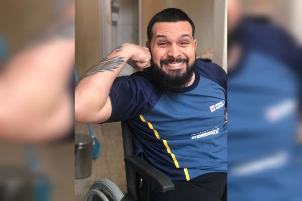 Foto mostra Erick Dias forçando os músculos e sorrindo sentado em sua cadeira de rodas