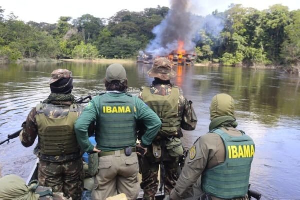 imagem colorida mostra agentes do ibama usando roupas militares verdes com colete a prova de balas em frente a rio onde balsa de garimpo ilegal queima - Metrópoles