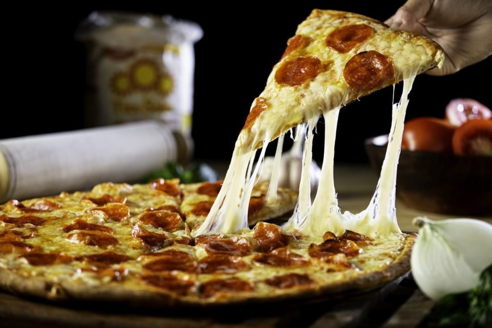 Foto colorida de pizza inteira com pedaço sendo levantado por uma mão e o queijo derretido ligando as fatias - Metrópoles