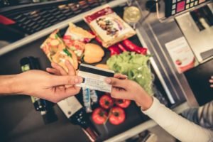 Cartão de vale-refeição sendo entregue na boca do caixa para um atendente de supermercado. Ao fundo, alimentos adquiridos na compra - Metrópoles