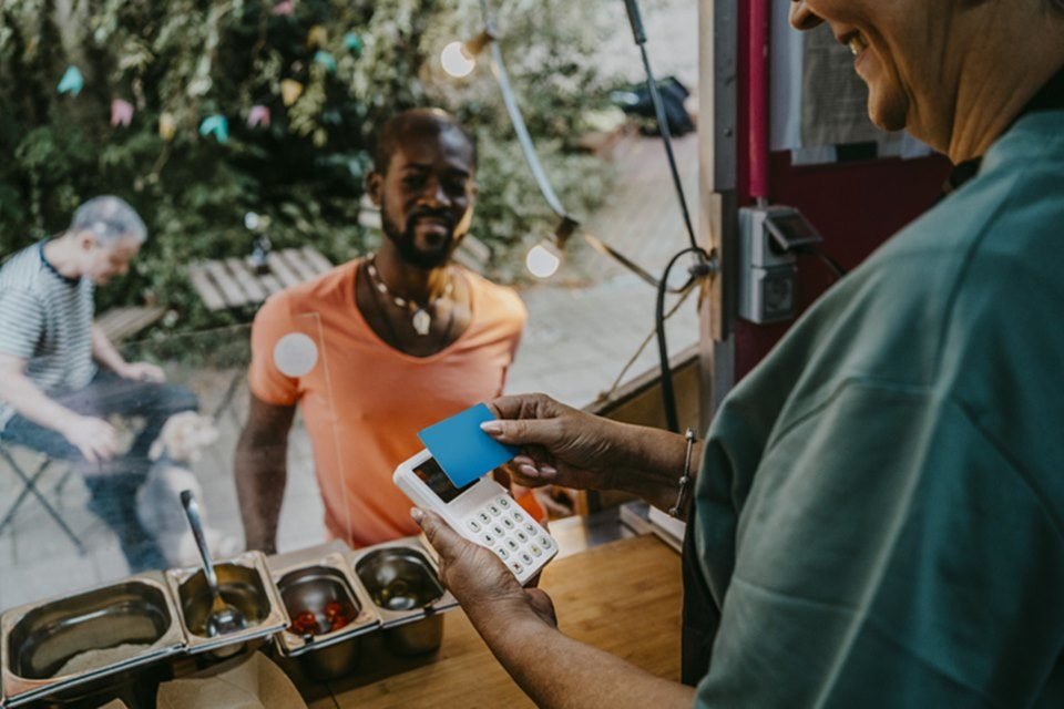 Imagem de homem com camiseta laranja fazendo pagamento com cartão em restaurante. Vendedor passa o cartão na maquininha - Metrópoles