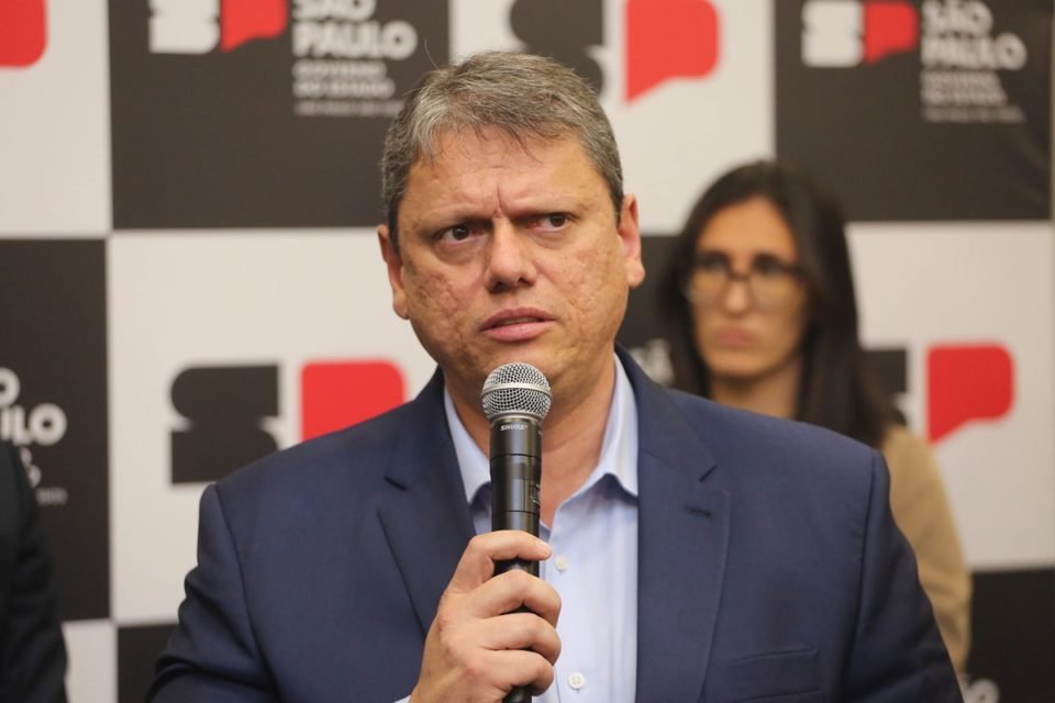 Imagem colorida mostra Tarcísio de Freitas, homem branco, grisalho e de terno azul, falando ao microfone à frente de um painel com logotipos do governo de SP - Metrópoles