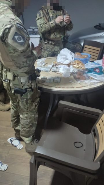 Policial com uniforme camuflado ao lado de mesa com diversos itens apreendidos em operação