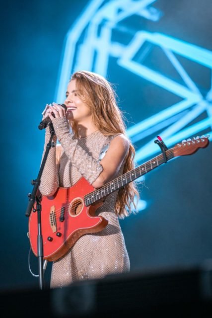 Foto do show de Giulia Be, realizado em Portugal. A cantora é branca, loira, está com vestido claro e toca uma guitarra - Metrópoles