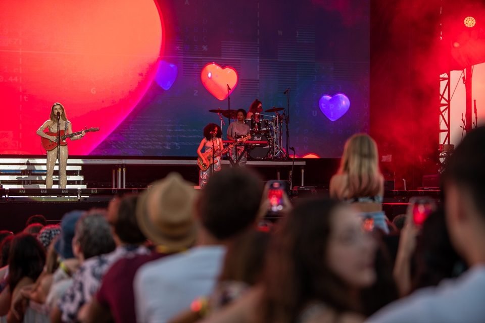 Foto do show de Giulia Be, realizado em Portugal. A cantora é branca, loira, está com vestido claro e toca uma guitarra - Metrópoles