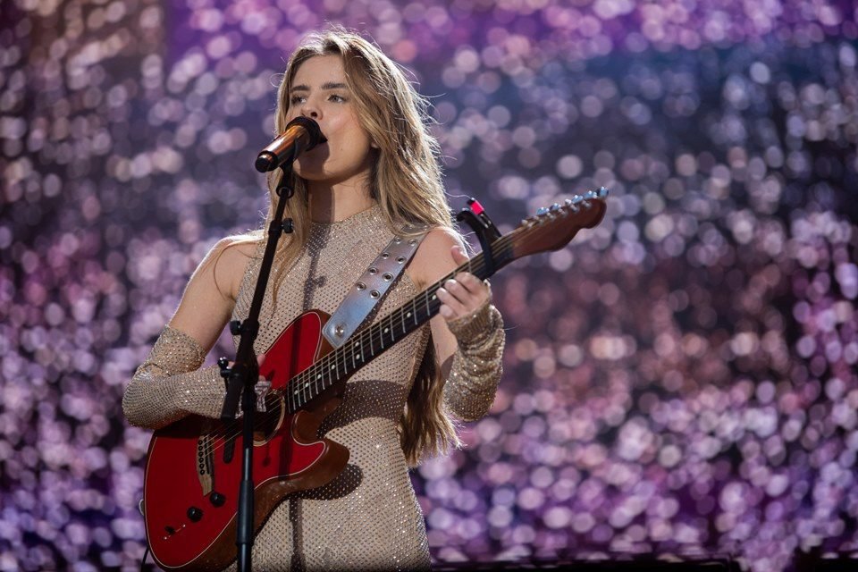 Foto do show de Giulia Be, realizado em Portugal. A cantora é branca, loira, está com vestido claro e toca uma guitarra - Metrópoles