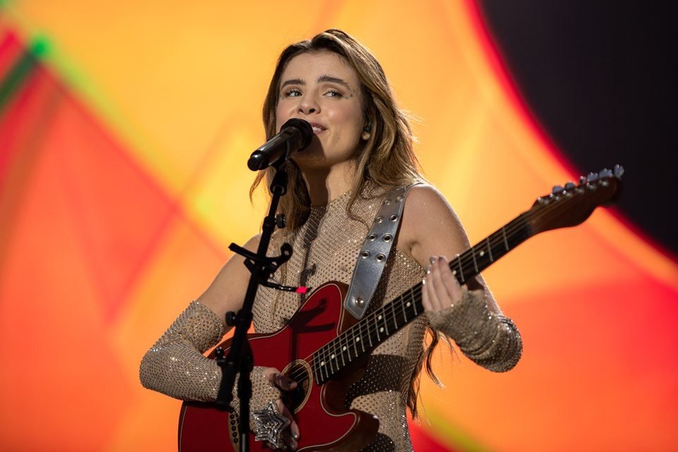 Foto do show de Giulia Be, realizado em Portugal. A cantora é branca, loira, está com vestido claro e toca uma guitarra - Metrópoles