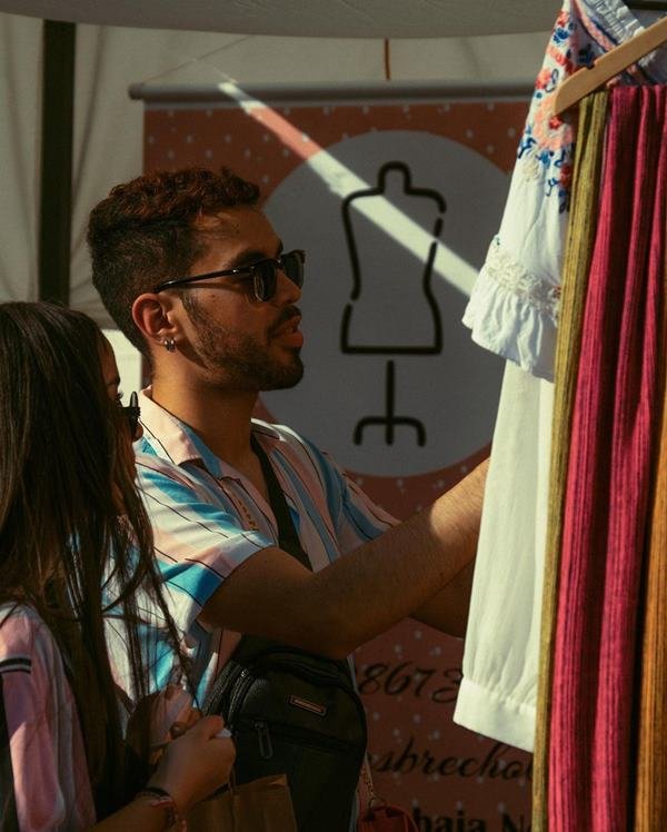 Cliente usando óculos de sol. Ele confere roupas em arara de uma feira - Metrópoles