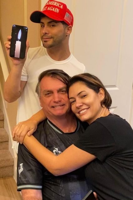 Michelle Bolsonaro Foto colorida de homem pardo, com boné vermelho e segurando um celular com a imagem de uma vela na tela. À frente, estão um homem idoso com blusa azul. No colo dele, tem uma mulher parda, com cabelo curto e blusa preta - Metrópoles