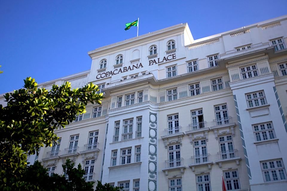 Imagem colorida da fachada do hotel Copacabana Palace, um dos ícones do Rio de Janeiro. Bandeira do Brasil tremula no topo do prédio, em dia de céu azul - Metrópoles