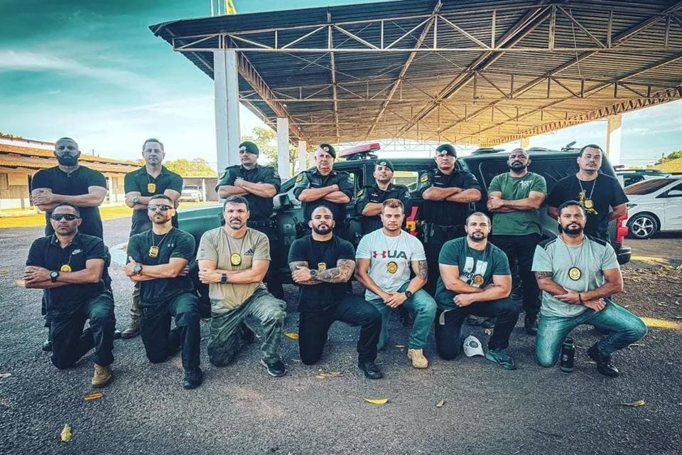 Homens agachados de joelhos, em frente a homens de braços cruzados, com viatura da polícia ao fundo - Metrópoles