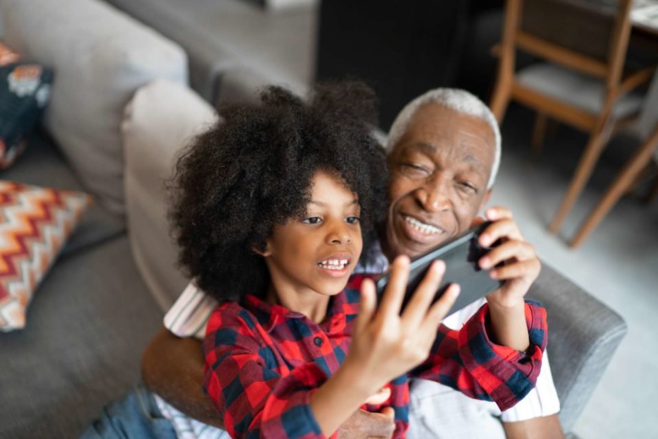 Foto mostra criança e idoso negros abraçados jogando juntos um jogo no telefone