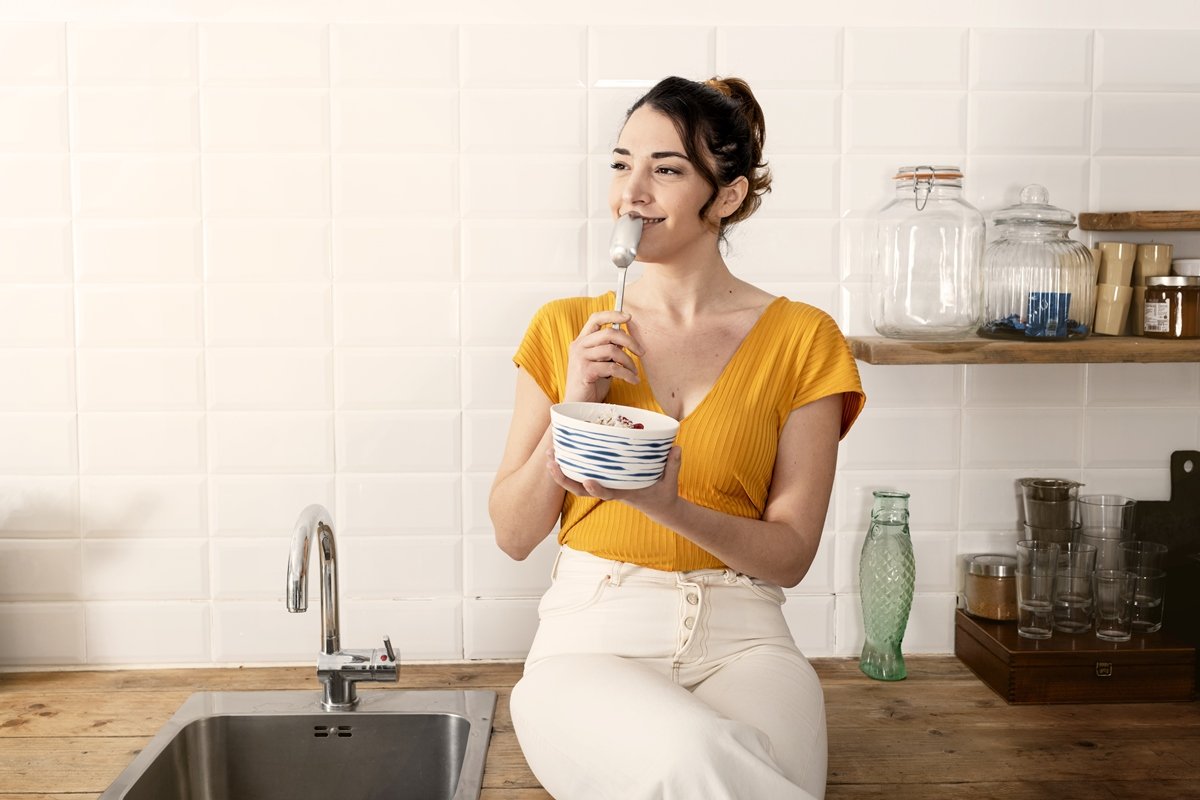Foto colorida. Mulher branca está encostada na bancada da cozinha segurando uma tigela com frutas e com a colher encostada nos lábios - Metrópoles