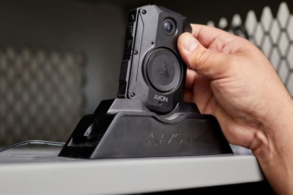 Imagem colorida da câmera usada no uniforme de PMs. Na foto, o dispositivo está sobre um suporte e uma mão segura a câmera - Metrópoles