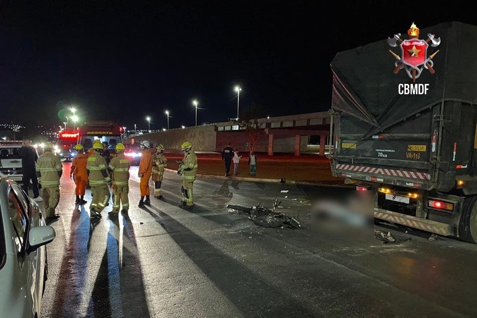 Cena de acidente de trânsito, com moto caída ao chão, atrás de caminhão com lona laranja parado na via; ao fundo, vê-se um viaduto; bombeiros militares uniformizados aparecem em pé ao lado da motocicleta