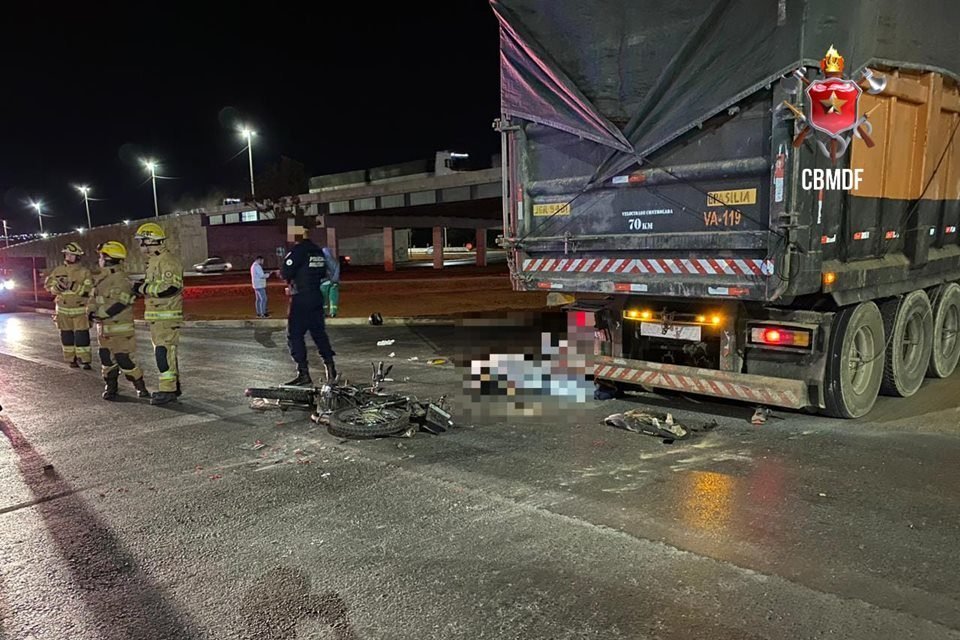 Cena de acidente de trânsito, com moto caída ao chão, atrás de caminhão com lona laranja parado na via; ao fundo, vê-se um viaduto; policial militar com uniforme azul aparece em pé ao lado da motocicleta