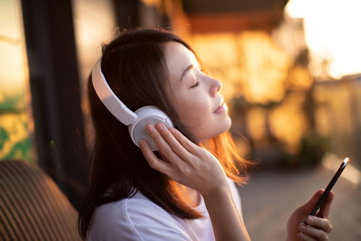 Foto colorida de mulher escutando música com mão em fone de ouvido - Metrópoles