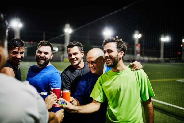 Foto mostra amigos próximos a campo de futebol fazendo brinde com cerveja