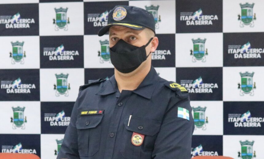 Em foto colorida GCM fardado usa máscara cirúrgica presa em frente a banner da corporação - Metrópoles