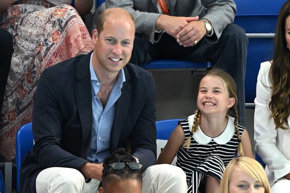 Foto colorida do príncipe William sentado ao lado da princesa Charlotte, os dois estão sorrindo - Metrópoles