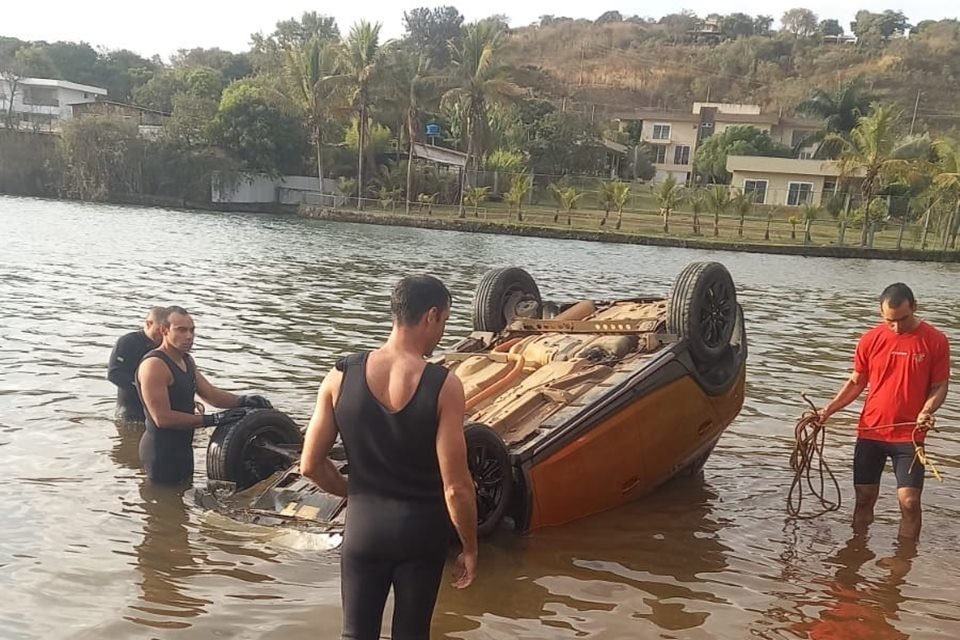 Imagem colorida de quatro homens dentro do Lago Paranoá, em Brasília, em torno de um carro laranja, com as rodas voltadas para cima, caído dentro da água; ao fundo, há casas, em área de vegetação rasteira