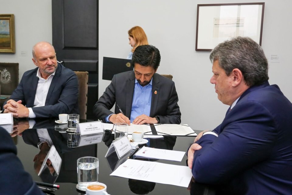 Imagem colorida mostra o prefeito Ricardo Nunes assinando um documento em uma mesa de reunião com o governador Tarcísio - Metrópoles