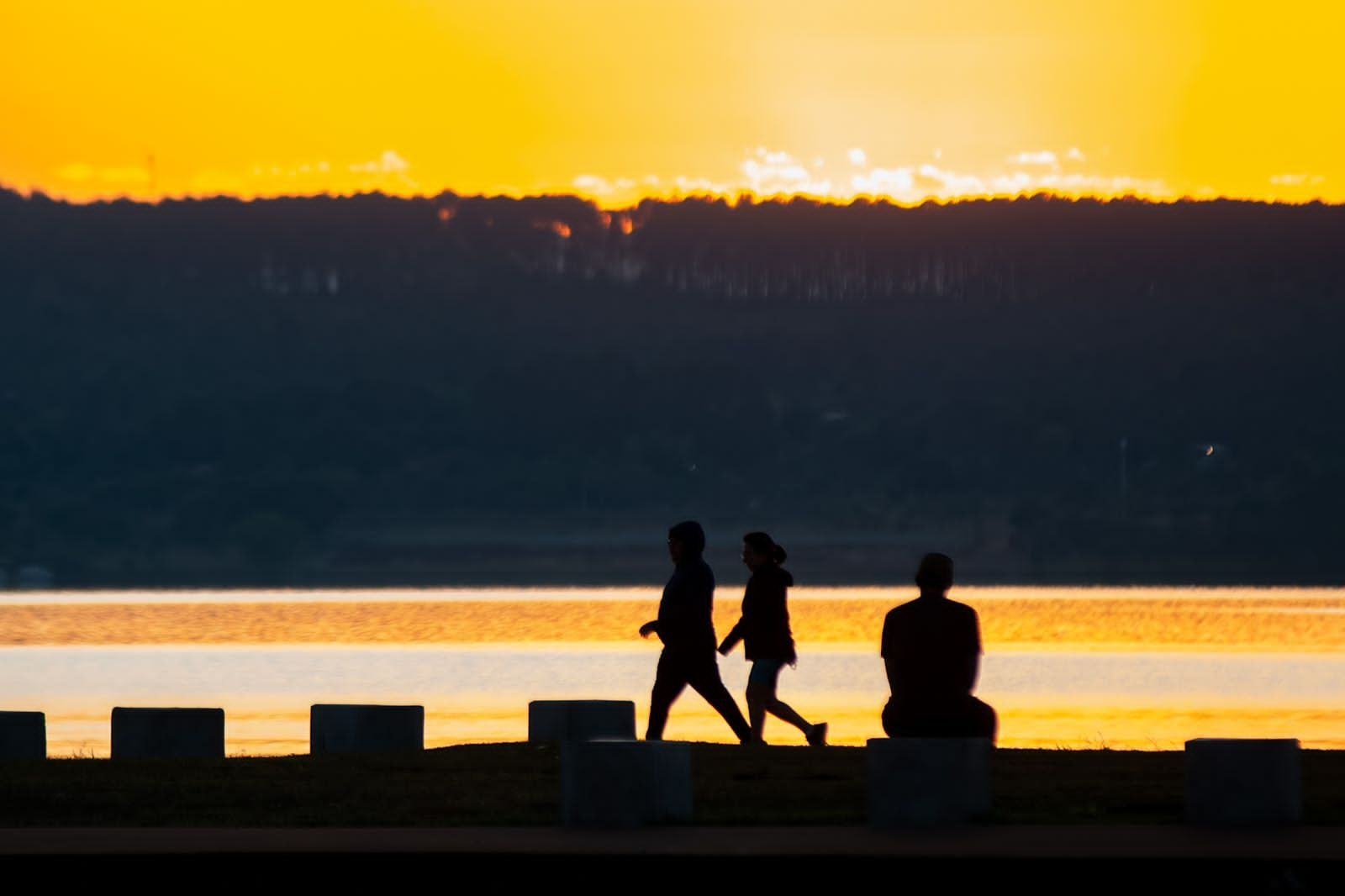 Pessoas caminham na orla do Lago Paranoá. O sol nasce ao fundo da imagem calor