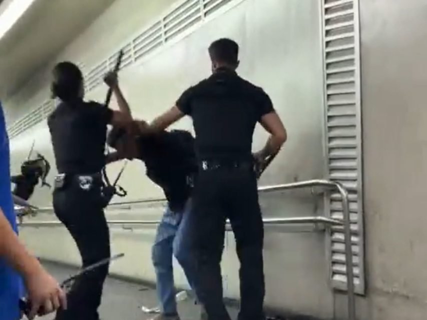 Imagem colorida mostra três funcionários do metrô, de preto, erguendo cacetetes para agredir um homem abaixado tentando se defender