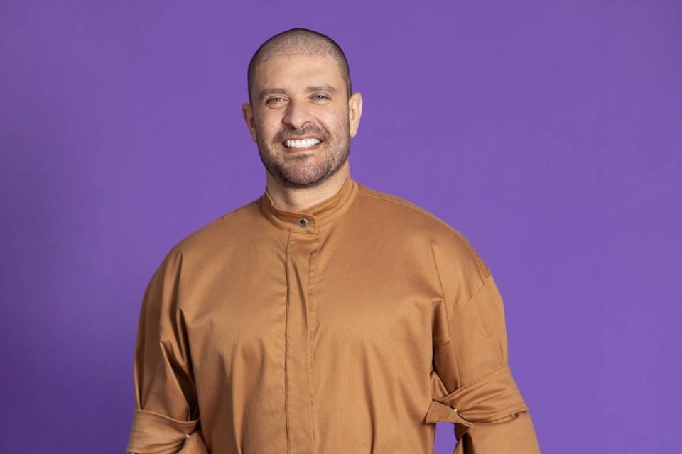 Cantor Diogo Nogueira vestido em macacão caramelo posa sorrindo em fundo roxo - metrópoles