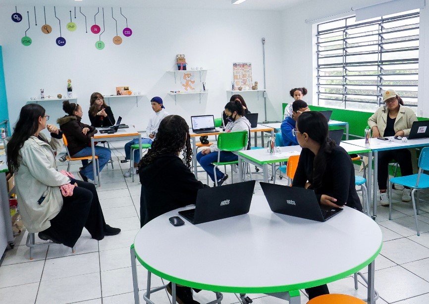 Foto colorida mostra professores e alunos conversando dentro uma sala de aula com paredes e mesas brancas - Metrópoles