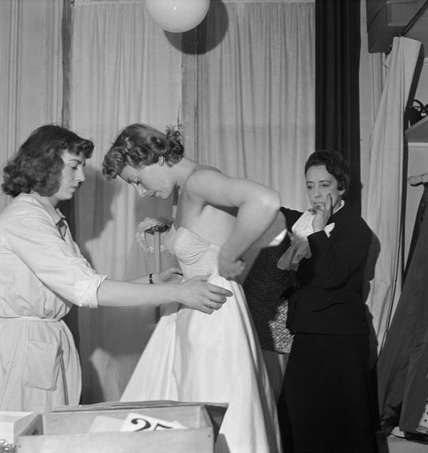 Na imagem em preto e branco, uma mulher prova um vestido, em ateli&ecirc; de moda, enquanto outra mulher ajuda e a estilista Elsa Schiaparelli observa - Metr&oacute;poles