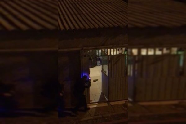 Homem com arma de choque ao lado de portão aberto à noite
