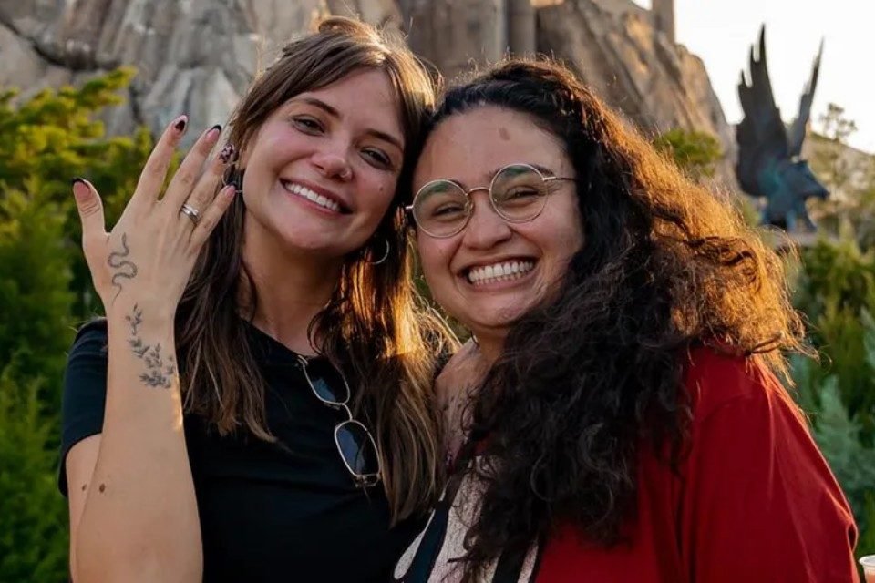 Foto colorida de Marcela McGowan e Luiza Martins. Elas estão abraçadas e Marcela mostra o anel de noivado. Ao fundo, uma montanha - Metrópoles