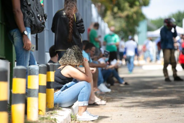 imagem colorida mostra pessoas sentadas no meio fio em entrada de faculdade para realizar prova de concurso da polícia federal - Metrópoles