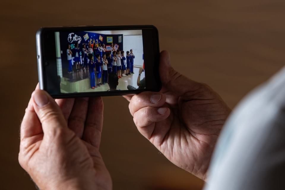 celular em primeiro plano com imagens de crianças nas escola. Celular é segurado por mãos de homem idoso